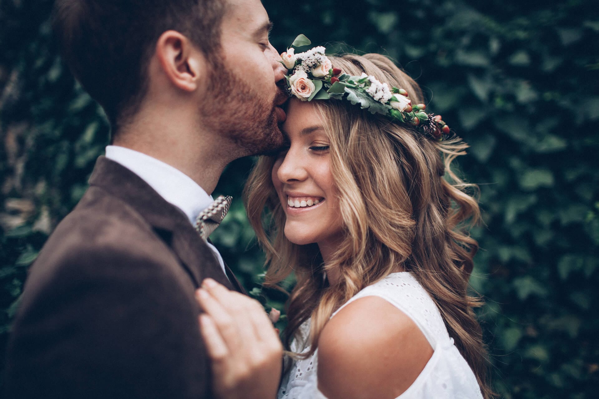 Groom kissing bride's forehead while she smiles, wearing a flower crown in an outdoor setting