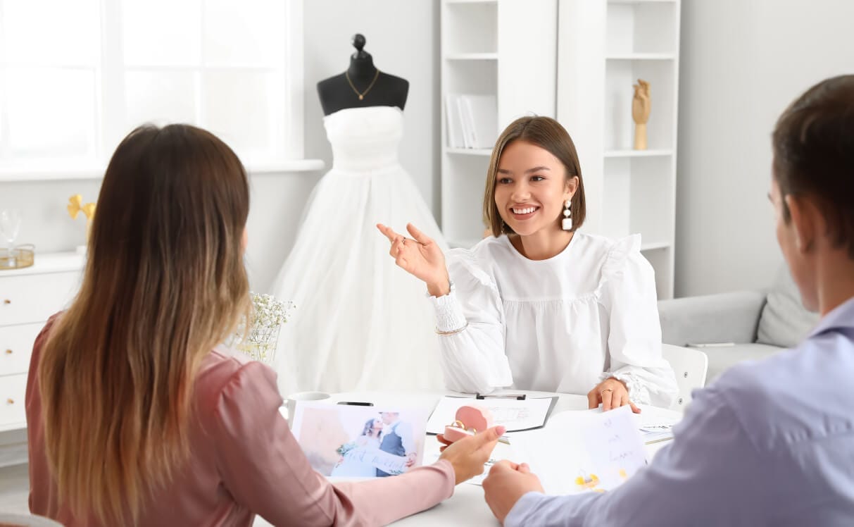 Wedding coordinator meeting with a couple in a bridal planning studio, with a wedding dress displayed in the background.