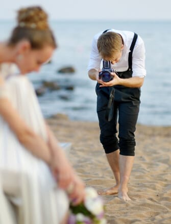 Photographer on a beach taking pictures of a bride, with the sea in the background.